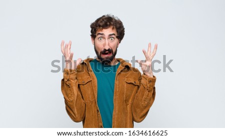 young handsome man screaming with hands up in the air, feeling furious, frustrated, stressed and upset against flat wall