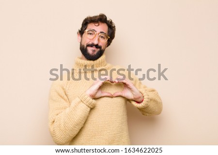 young cool man smiling and feeling happy, cute, romantic and in love, making heart shape with both hands against flat wall