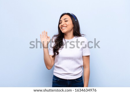young latin pretty woman smiling happily and cheerfully, waving hand, welcoming and greeting you, or saying goodbye against flat wall