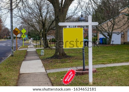 Blank yellow sign on a white post on a lawn in the front yard of a house near a sidewalk and a street