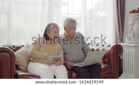 Portrait asian senior couple sitting on coach, elderly man using laptop and senior woman use tablet, They are discussing together with happy