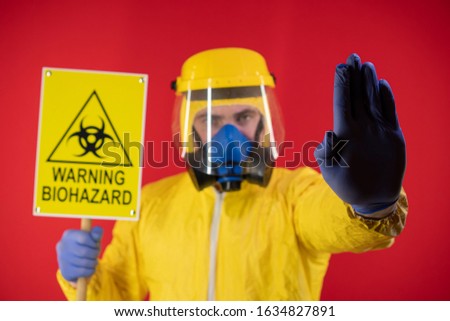 A man in a protective chemical mask and a protective yellow suit with a banner in his hand with the words biohazard shows a stop sign. Biohazard concept