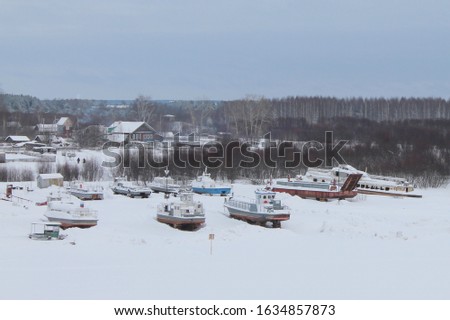 Snow covered landing stage with ships on background of wooden houses and forest. Frozen river surface. Cloudy winter day. Stock photo for web, print, background and wallpaper with epty space for text.