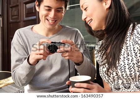 Joyful Japanese tourist couple sitting at a cafe terrace shop in London city, using a consumer digital photo camera to look at their trip pictures, smiling outdoors during a sunny day.