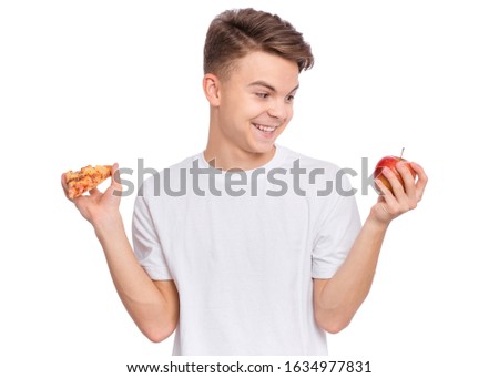 Happy handsome young teen boy choosing slice of pizza instead of red apple. Close up portrait of cute child with healthy and unhealthy food, isolated on white background. Guy looks at apple.
