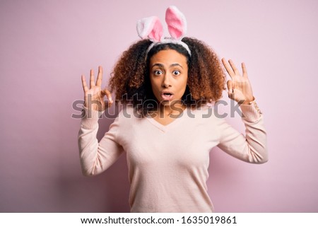 Young african american woman with afro hair wearing bunny ears over pink background looking surprised and shocked doing ok approval symbol with fingers. Crazy expression