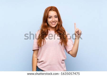 Teenager redhead girl over isolated blue background showing and lifting a finger in sign of the best