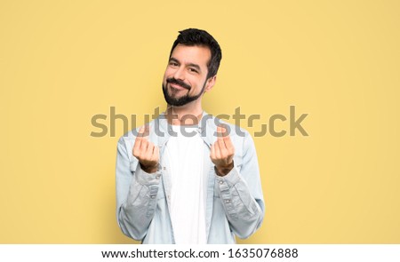 Handsome man with beard making money gesture over isolated yellow background