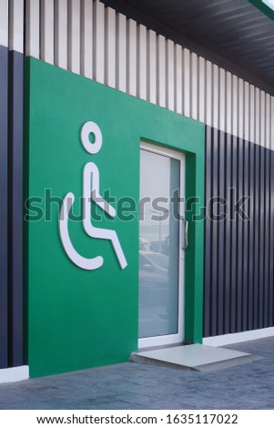 White disabled wheelchair  sign with glass door on green wall and wooden panel wall decorations in public restroom area, side view and vertical frame 