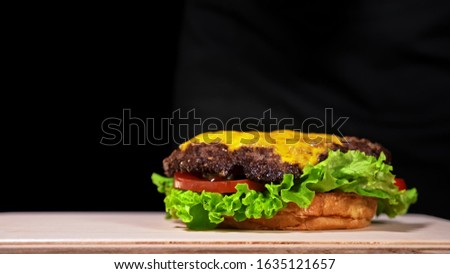 Craft burger is cooking on black background in black food gloves. Consist: red sauce salsa, lettuce, red onion, pickle, cheese, chilli green pepper, air bun and marbled meat beef. Not made ideal