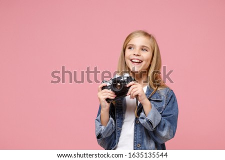 Laughing little blonde kid girl 12-13 years old in denim jacket posing isolated on pastel pink background in studio. Childhood lifestyle concept. Mock up copy space. Hold retro vintage photo camera