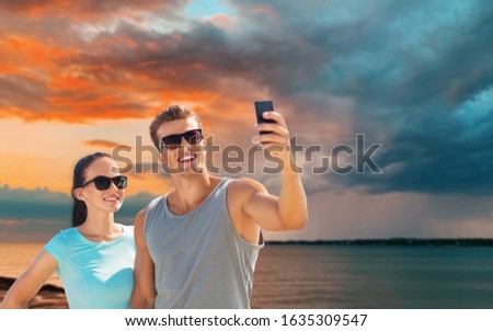 fitness, sport and healthy lifestyle concept - happy couple in sports clothes and sunglasses taking selfie by smartphone over sea and sunset sky background