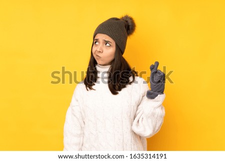 Young woman with winter hat over isolated yellow background with fingers crossing and wishing the best