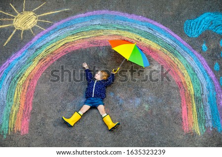 Happy little toddler girl in rubber boots with rainbow sun and clouds with rain painted with colorful chalks on ground or asphalt in summer. Cute child with umbrella having fun. creative leisure