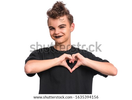Portrait of crazy teen boy with spooking make-up showing heart symbol and shape with hands. Young teenager in style of punk goth dressed in black making heart, isolated on white background. 