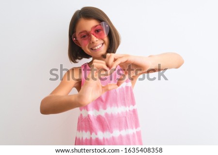 Young beautiful child girl wearing pink t-shirt and sunglasses over isolated white background smiling in love showing heart symbol and shape with hands. Romantic concept.
