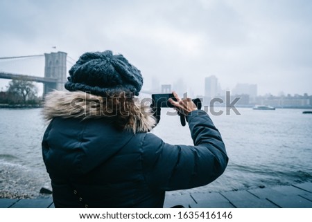 Tourist woman with warm clothes takingaphoto of New York Skyline from DUMBO neighborhood, while sightseeing new york during winter season.