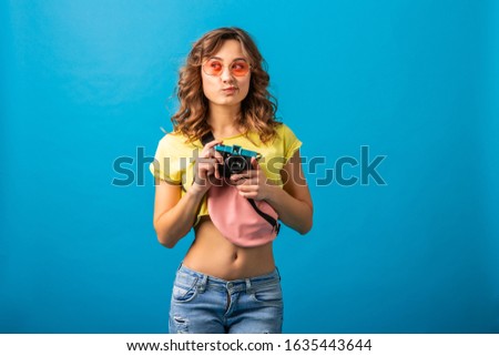 attractive thinking woman posing with vintage photo camera taking pictures dressed in hipster summer colorful outfit isolated on blue studio background, traveler photographer hobby