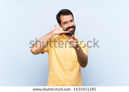 Young man with beard over isolated blue background making phone gesture and pointing front