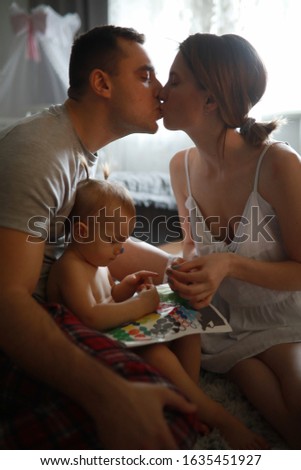 Baby girl sits and plays next to kissing parents.