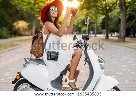 attractive smiling woman riding on motorbike in street in summer style outfit wearing white dress and red hat traveling on vacation, taking pictures on vintage photo camera