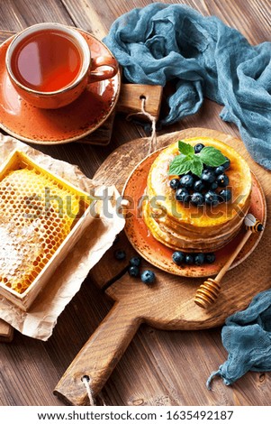 Pile of small homemade pancakes with berries, tea cup and honeycomb on wooden table