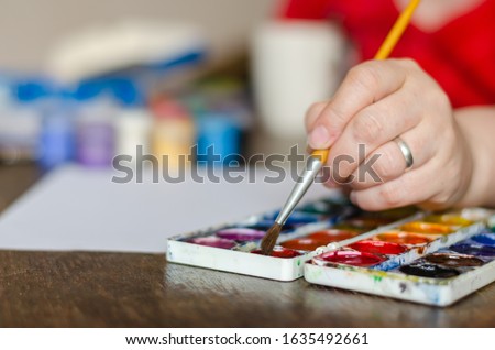 Selective focus of a female hand soaking brush in watercolor paint. A married woman is engaged in creativity sitting at a table. Eye level shooting. Close-up.