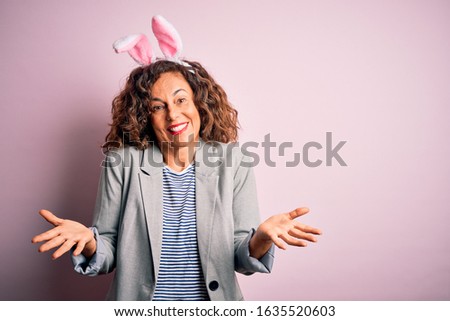 Middle age beautiful woman wearing bunny ears standing over isolated pink background clueless and confused expression with arms and hands raised. Doubt concept.