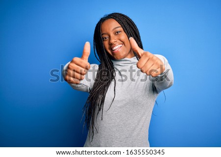 Young african american woman standing wearing casual turtleneck over blue isolated background approving doing positive gesture with hand, thumbs up smiling and happy for success. Winner gesture.
