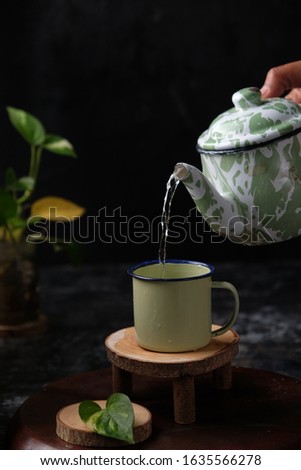 Pouring water from vintage green enamel teapot into an enamel cup on wooden coaster with leaves. dark  background