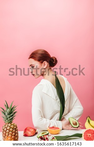Pensive ginger woman with pineapple, bananas, garnets and kiwi. Studio shot of attractive girl with tropical fruits on pink background.