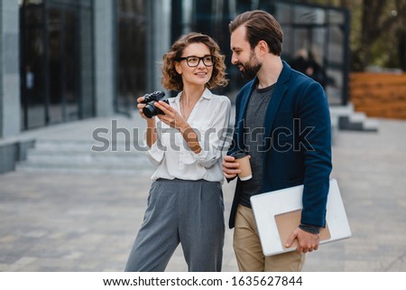 man and woman talking in urban city center in business style working together, holding laptop, smiling, stylish freelance people, discussing, taking pictures for project on digital photo camera