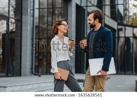 attractive couple of man and woman talking in urban city center in smart casual business style working together, holding laptop, smiling, stylish freelance people, discussing, drinking coffee