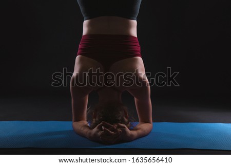 Beautiful young woman working out in a studio on black background, doing yoga exercise on blue mat, variation of supported headstand, garuda salamba sirsasana.