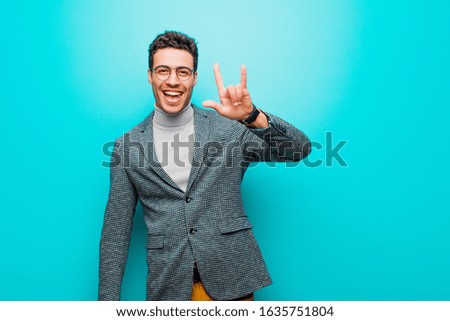 young arabian man feeling happy, fun, confident, positive and rebellious, making rock or heavy metal sign with hand against blue wall