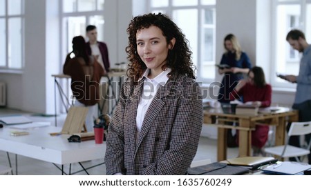 Beautiful portrait of young European business woman touching curly hair in formal suit, smiling happy at modern office.