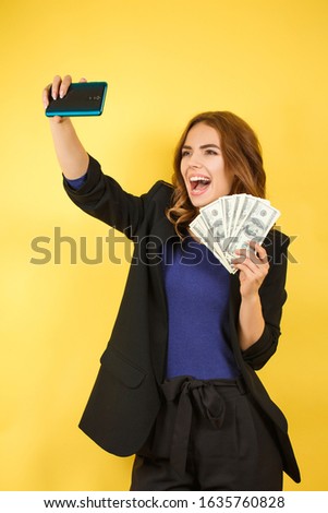Happy woman in a jacket makes selfie with money, on a yellow background, good earnings. freelancer business lady concept. successful purchase and money saved