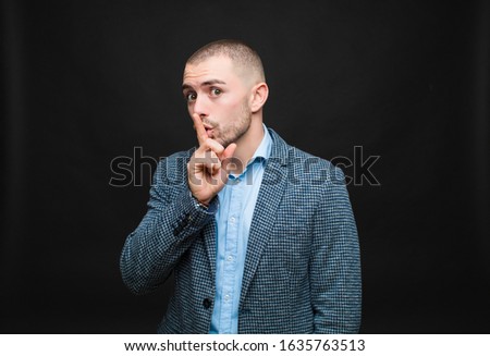 young businessman asking for silence and quiet, gesturing with finger in front of mouth, saying shh or keeping a secret against flat wall