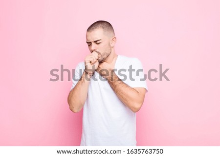 young handsome man feeling ill with a sore throat and flu symptoms, coughing with mouth covered against flat wall