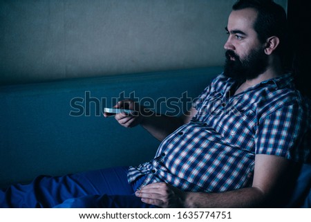Laziness, sedentariness, overweight, daily routine, television addiction. Man sitting at coach watching TV late at night