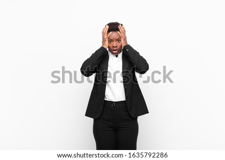 young pretty black womanfeeling horrified and shocked, raising hands to head and panicking at a mistake against white wall