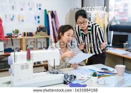 two asian chinese women meeting in fashion design studio. female creative dressmakers talking about plan on sketch paper at garment factory. lady colleagues discussing design of new dress on painting