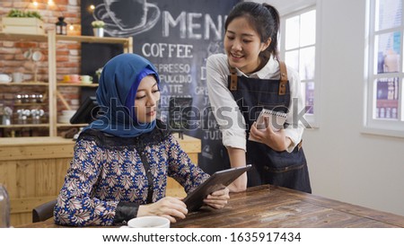 Portrait of smiling young japanese waitress taking order from malay client in cafe bar writing note. muslim woman guest using digital tablet reading menu while friendly female barista introduce
