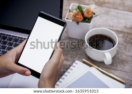 Mockup image of woman hand holding and using black mobile smart phone with blank white screen, work on laptop computer with tablet and cup of coffee on wooden table at home .