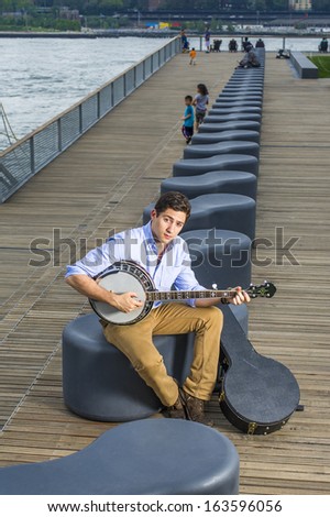 Dressing in a light blue shirt, dark yellow jeans and brown boot shoes, a young musician with an instrument box is sitting on a modern style bench, playing a banjo. / Play music Outside