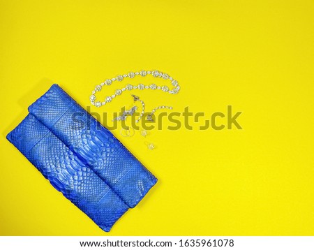 Top view of navy python clutch with woman's jewellery; earring, necklace, hairpin, rings, lay flat on yellow background with copy space. For fashion and beauty theme.