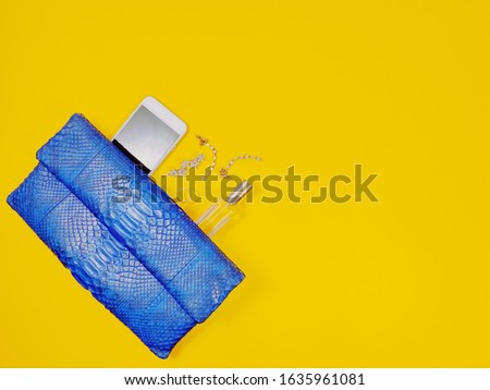 Top view of navy python clutch with woman's jewellery; earring, necklace, hairpin, rings, cell phone, lay flat on yellow background with copy space. For fashion and beauty theme.