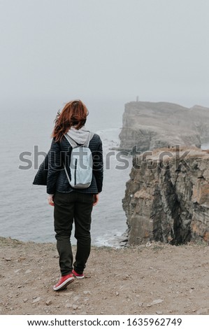 young brunette girl with a backpack stands with her back and watches the gray sea and sheer cliffs in the fog of the island, a postcard about travel and discoveries of the world