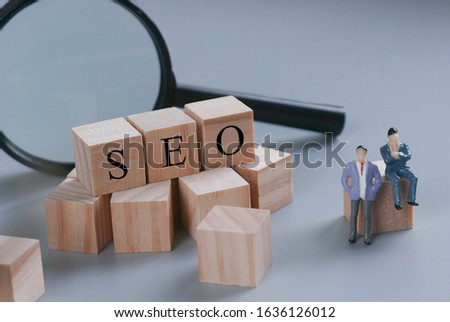 Minoature people with magnifying glass and wooden blocks written with SEO (search engine optimization) on grey background.