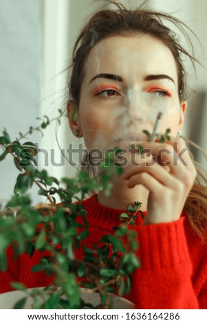 
Portrait of a young woman with pink make-up and career eyes, with a tail on her head in a red sweater that holds a green plant in a pot and smokes a cigarette